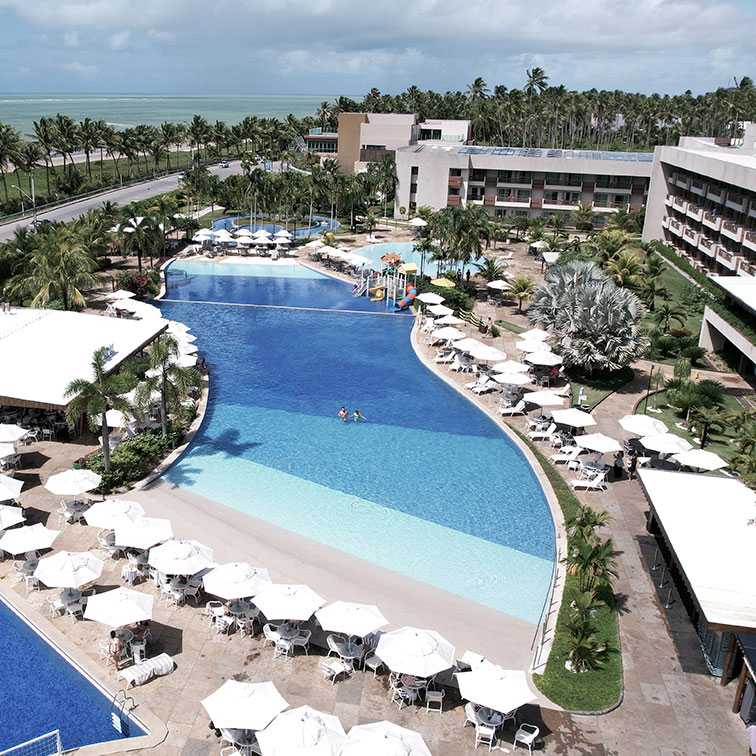 Vista aérea do resort destacando as piscinas do Verano, sendo uma com faixa de areia artificial. Tem vários guarda-sóis e espreguiçadeiras brancas ao redor da piscina, coqueiros, estrutura do Japaratinga Lounge Resoort e a vista do mar e da praia ao fundo.