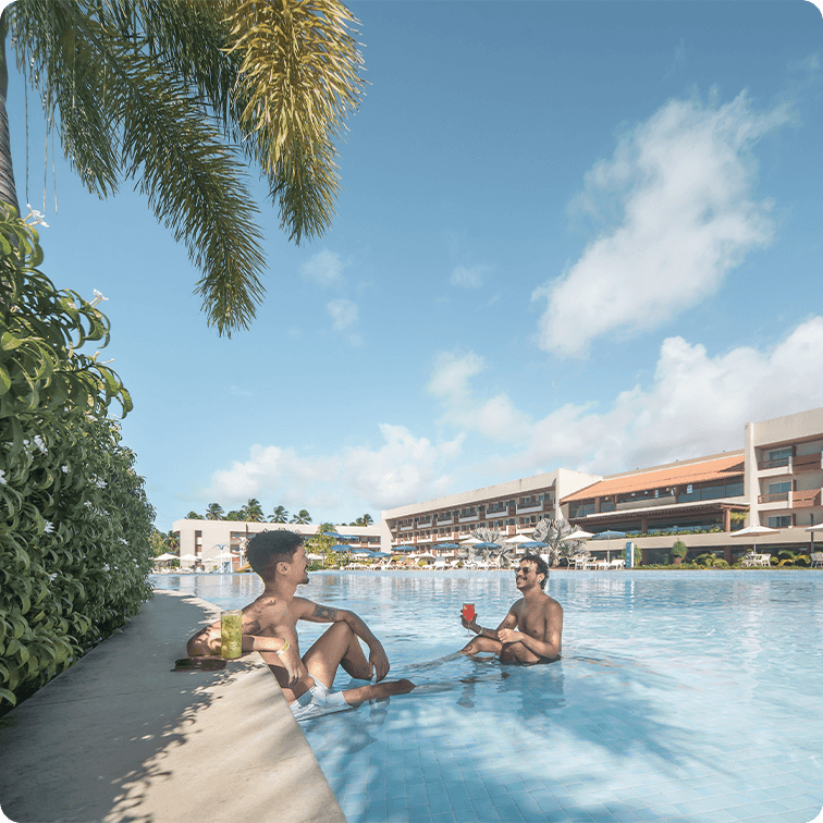 Dois jovens em uma piscina do Japaratinga Lounge Resort, um sentado na borda e outro parcialmente imerso, ambos sorrindo e segurando drinks coloridos em um dia de sol. O fundo mostra a estrutura do resort e vegetação tropical.
