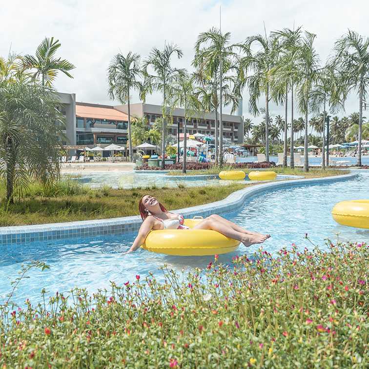 Mulher deitada e sorrindo, flutuando em uma boia amarela no rio lento do Japaratinga Lounge Resort. A água é azul-clara e a área é cercada por vegetação tropical, coqueiros e o prédio do resort ao fundo.
