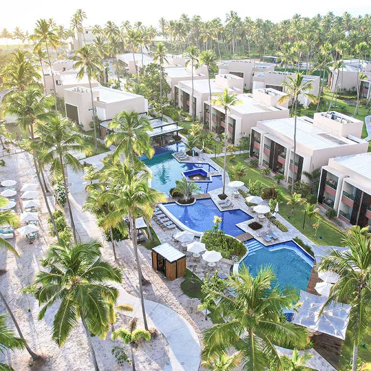 Vista aérea do Japaratinga Lounge Resort com a visão das novas acomodações Garden, cercado por um denso coqueiral. No centro, a área de piscinas da nova expansão com algumas piscinas, espreguiçadeiras, mesas com guarda-sol e o Mangô Bar. 
