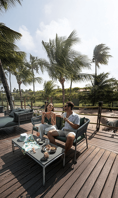 Casal sorrindo e relaxando em confortáveis sofás de área externa no deck de madeira de um resort. Eles estão com petiscos e bebidas, com a mulher segurando um espetinho de camarão. A cena é sombreada por coqueiros e rodeada por vegetação tropical.