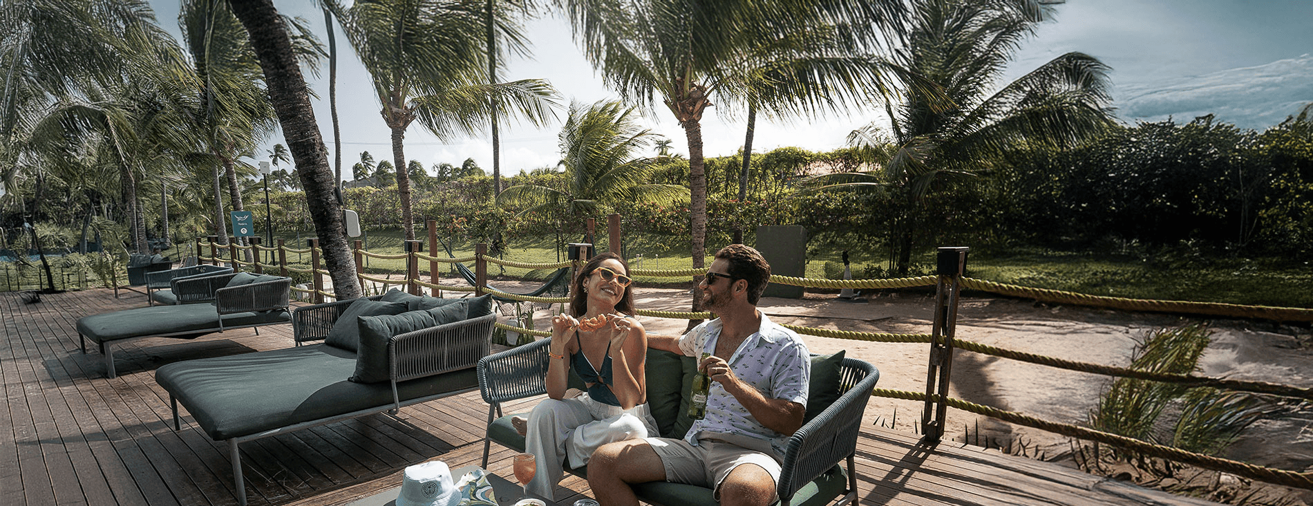 Casal sorrindo e relaxando em confortáveis sofás de área externa no deck de madeira de um resort. Eles estão com petiscos e bebidas, com a mulher segurando um espetinho de camarão. A cena é sombreada por coqueiros e rodeada por vegetação tropical.