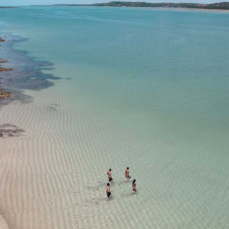 Vista aérea da Croa de São Bento, com a maré baixa revelando um banco de areia e áreas de recifes de corais expostos. Quatro pessoas estão caminhando na água rasa e transparente, com o mar azul-claro se estende até o horizonte.