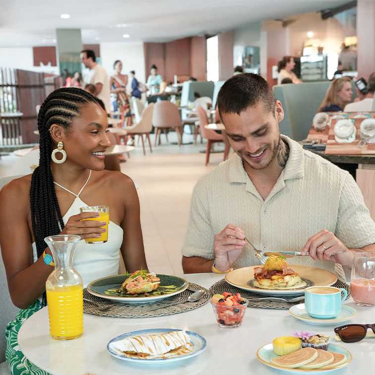 Um casal, sorrindo e desfrutando de um café da manhã na mesa do restaurante, com várias opções de pratos (tapioca, panquecas, frutas, ovos) e sucos. O ambiente é claro, moderno e movimentado.