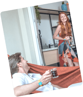 Casal aproveitando a Garden Exclusive, categoria do Japaratinga Lounge Resort, ele deitado na rede na varanda com um café na mão e ela em pé também com café na mão, com vestido longo.