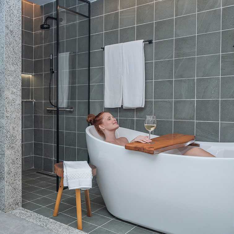 Uma mulher ruiva relaxando na acomodação Garden Exclusive, em uma banheira moderna e branca, com uma taça de vinho branco em uma bandeja de madeira sobre a banheira. Ao lado há um chuveiro com box de vidro e paredes de azulejo cinza, e uma planta verde em um vaso.