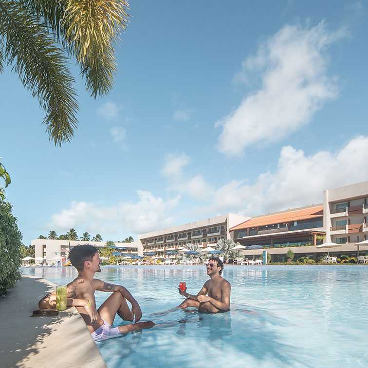 Dois jovens em uma piscina do Japaratinga Lounge Resort, um sentado na borda e outro parcialmente imerso, ambos sorrindo e segurando drinks coloridos em um dia de sol. O fundo mostra a estrutura do resort e vegetação tropical.