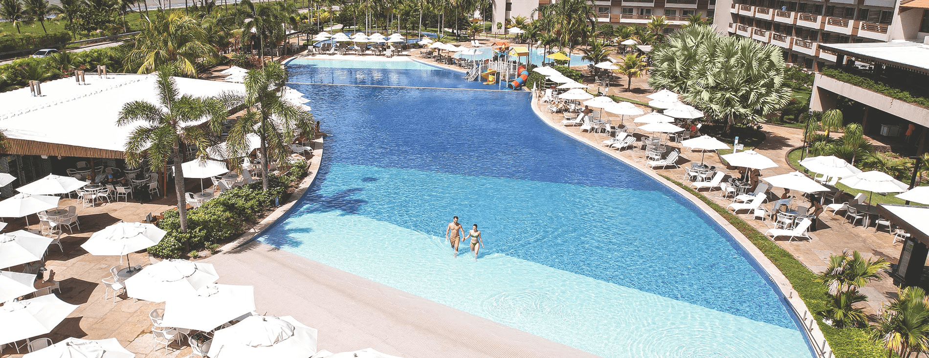 Vista aérea da piscina principal. O entorno é repleto de guarda-sóis e mesas brancas, coqueiros e o edifício principal do resort. Ao fundo, o mar azul com a linha de coqueiros na praia sob um céu parcialmente nublado.