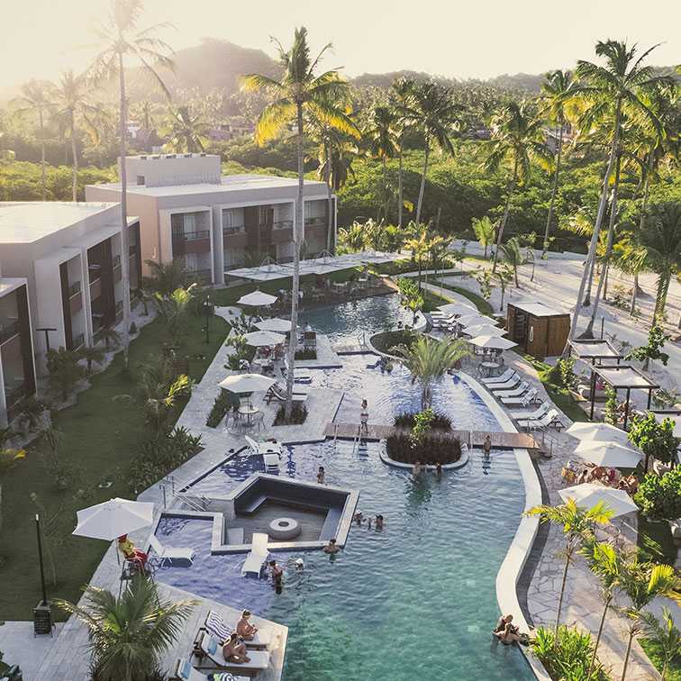 Vista aérea do resort com coqueiros densos. A área central de piscinas de formato orgânico ao lado do Garden e pessoas aproveitando a piscina. Tem alguns bangalôs, guarda sol na mesa e espreguiçadeiras na beira da piscina. A cena é banhada por uma luz dourada intensa de fim de tarde ou nascer do sol.