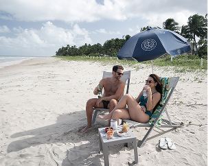 Casal conversando e sorrindo no apoio de praia do Japaratinga Lounge Resort. Eles estão com bebidas nas mãos e com petiscos na mesa.