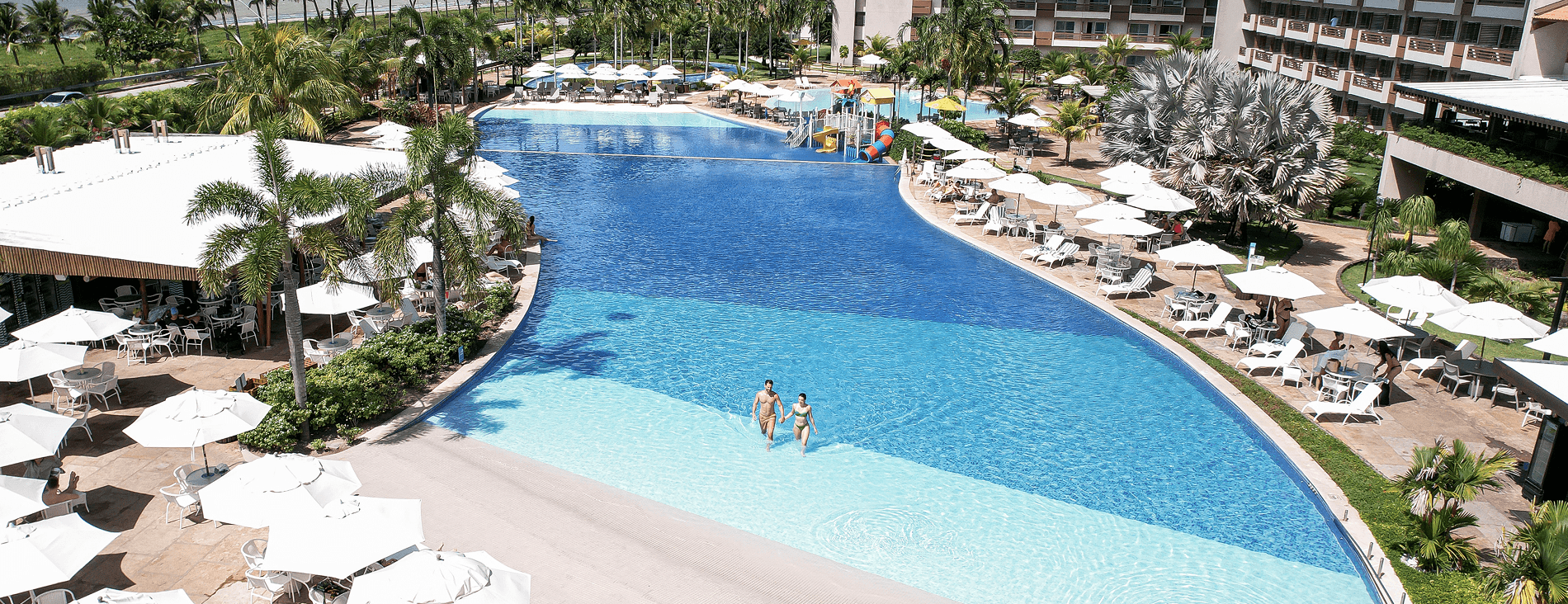 black friday japaratinga lounge resort Vista aérea da piscina principal. O entorno é repleto de guarda-sóis e mesas brancas, coqueiros e o edifício principal do resort. Ao fundo, o mar azul com a linha de coqueiros na praia sob um céu parcialmente nublado.