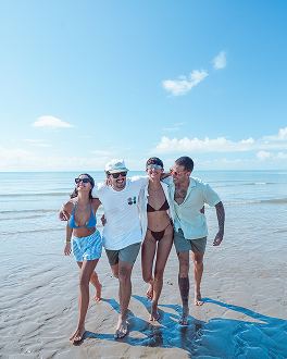 Quatro amigos abraçados enquanto caminham na beira mar com o oceano e o céu azul ao fundo.