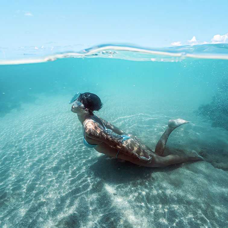 Mulher jovem em um biquíni azul-claro fazendo snorkeling e mergulhando sob a superfície em águas azul-turquesa e cristalinas. O fundo de areia clara é visível e o céu azul aparece acima da linha d'água.