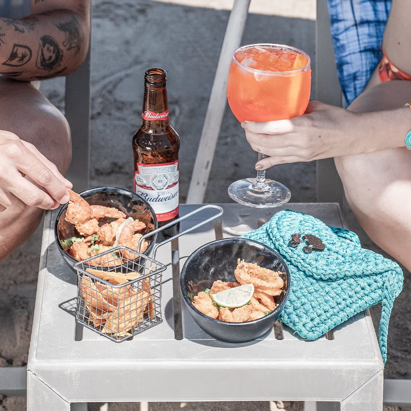 Mesa na areia com cerveja, drink alaranjado e porções de petiscos diversos, com um casal ao redor.