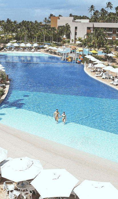 Vista aérea da piscina principal. O entorno é repleto de guarda-sóis e mesas brancas, coqueiros e o edifício principal do resort. Ao fundo, o mar azul com a linha de coqueiros na praia sob um céu parcialmente nublado.