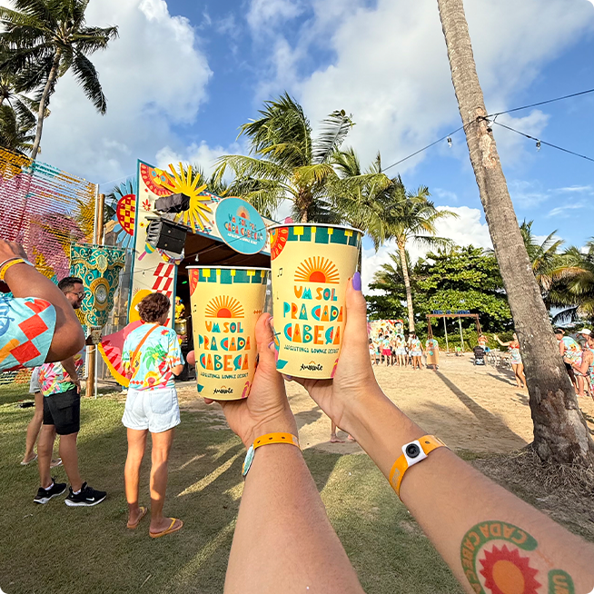 Multidão de pessoas com abadás temáticos dançando em frente ao palco colorido com banda ao vivo. O palco tem o tema um sol pra cada cabeça e está cercado por coqueiros.