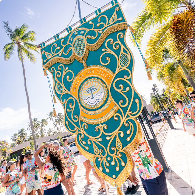Um estandarte grande, ornamentado e colorido sendo carregado pelo bloco de carnaval ao ar livre. A cena mostra uma multidão abadá, com coqueiros ao fundo em um dia ensolarado.