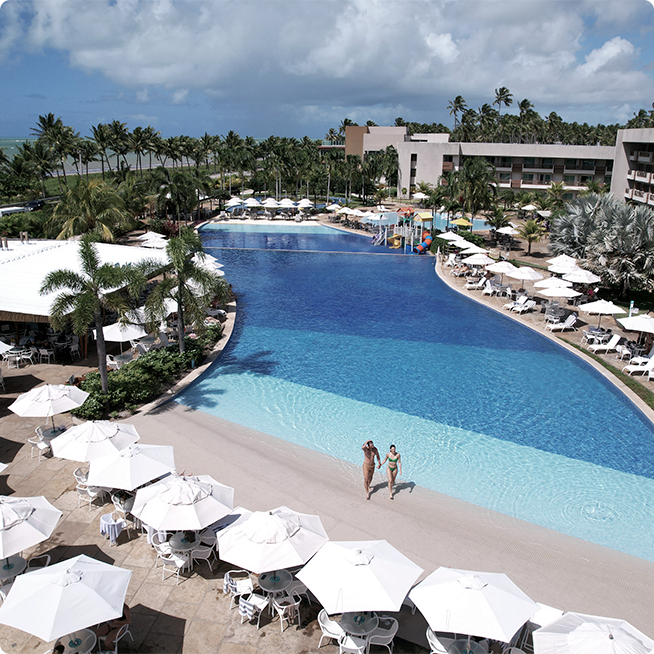 Vista aérea da piscina principal do Japaratinga Lounge Resort. A piscina possui uma praia artificial, cercada por alguns guarda-sóis brancos e vegetação tropical.