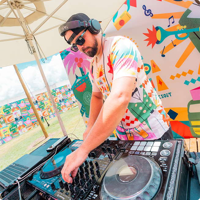 DJ tocando música na festa de carnaval com um guarda-sol protegendo. Vestido com o abadá oficial e um backdrop também na mesma estampa.