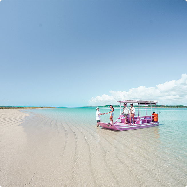Barco com grupo de amigos no raso da Croa de São Bento, com água em tom turquesa e céu sem nuvens. Ao fundo se vê a vegetação de Alagoas.