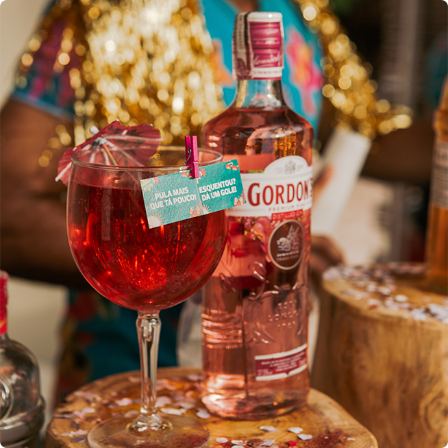 Gin Gordon cor de rosa, com taça cheia em cima de um toco de madeira, e o bartender ao fundo servindo outras bebidas.
