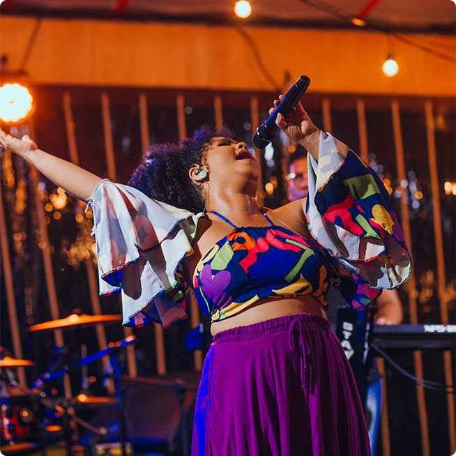 Cantora no palco do Japaratinga Lounge Resort cantando com banda atrás, instrumentos como teclado e bateria são visíveis.