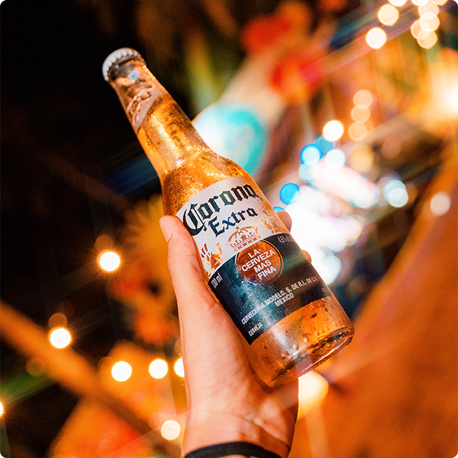 Cerveja Corona na mão de uma pessoa, há luzes, palmeiras e o palco do carnaval com fundo desfocado.