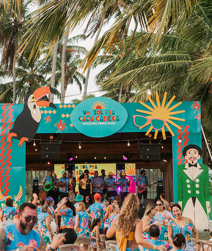 Palco de show de carnaval com letreiro 'Um sol pra cada cabeça' com banda ao vivo, multidão de pessoas dançando e vestindo abadás temáticos. Cenário de praia com coqueiros.