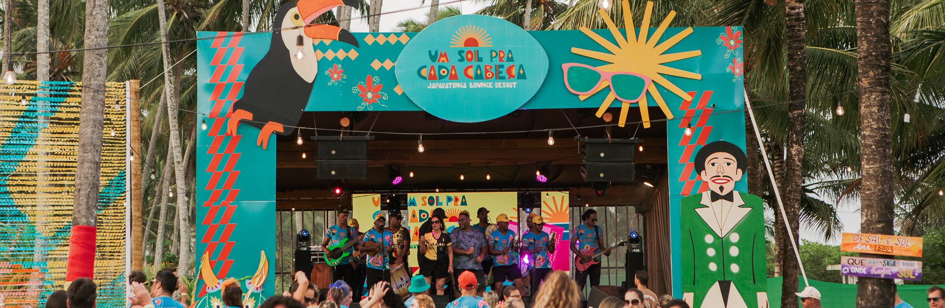 Palco de show de carnaval com letreiro 'Um sol pra cada cabeça' com banda ao vivo, multidão de pessoas dançando e vestindo abadás temáticos. Cenário de praia com coqueiros.
