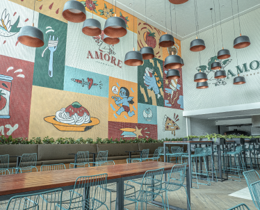 A imagem mostra o interior vibrante de um restaurante contemporâneo, com um mural artístico colorido e temático que adorna a parede. Ele apresenta elementos gráficos relacionados à comida e à cozinha italiana, incluindo a palavra "AMORE", nome da forneria, e imagens estilizadas de tomates, pizza e outros itens culinários, todos em um estilo de arte pop expressivo. Abaixo há longas mesas de madeira e cadeiras com detalhes em azul turquesa. Acima das mesas, luminárias modernas com tons de cobre criam uma iluminação aconchegante.