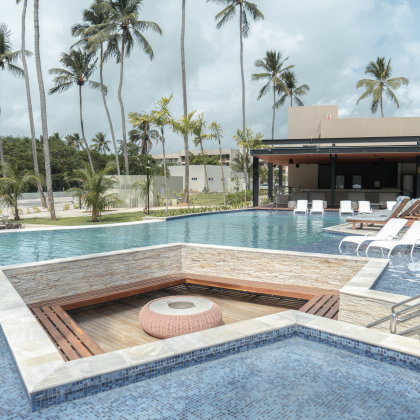 Piscina de águas cristalinas em um resort, cercada por um cenário de palmeiras altas e céu nublado. O destaque vai para uma jacuzzi embutida com assentos de madeira e um confortável puff redondo no centro, prometendo relaxamento total. Espreguiçadeiras brancas estão dispostas ao longo da borda da piscina.