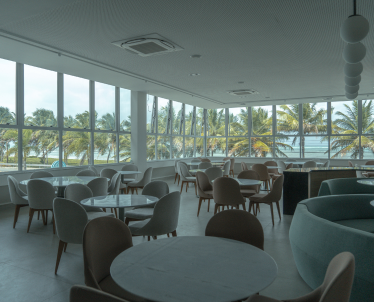 A fotografia mostra o interior de um restaurante elegante dentro de um resort, com uma ampla janela que oferece uma vista panorâmica das palmeiras e do oceano turquesa ao fundo. Cadeiras e mesas de centro modernas de madeira com tampo de mármore estão arranjadas de maneira convidativa, enquanto sofás circulares de tecido azul acrescentam um toque de cor e conforto.