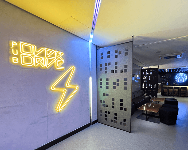 Entrada do Pub Overdrive no Japaratinga Lounge Resort. Com letreiro do Pub em amarelo com grande raio luminoso. Ao fundo tem a logo no Japaratinga Lounge Resort luminoso em cor azul, e o ambiente com mesas e cadeiras.