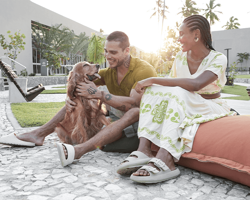 Casal se divertindo com seu cachorro na Praça de Coqueiros no Japaratinga Loungue Resort no fim de tarde.