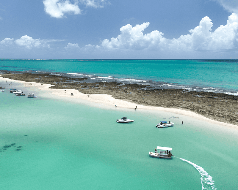 Vista aérea da Croa de São Bento com recifes de coral expostos pela maré baixa, formando piscinas naturais. A água é de um azul-turquesa transparente, com algumas pessoas nadando no mar, e algumas embarcações ancoradas na faixa de areia, o céu esta sem nuvens.