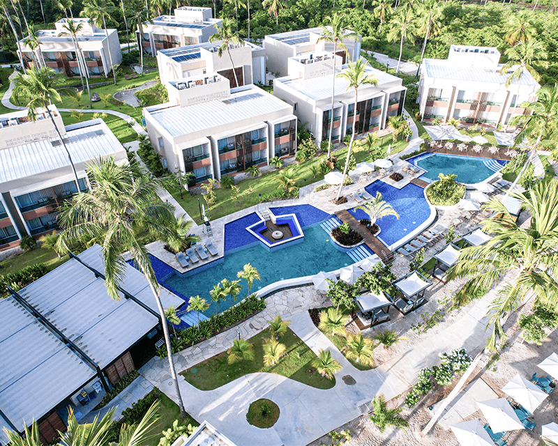 Vista aérea das Gardens Suítes do Japaratinga Lounge Resort, mostrando a área das piscinas com guarda-sóis brancos, espreguiçadeiras, cercadas por vegetação tropical e palmeiras