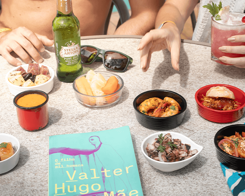 Diversos petiscos como camarão, queijos, frutas, polvo picado, entre outros e bebidas como a cerveja Stella Artois e um drink vermelho. As mãos de um casal está sob a mesa, que há também um livro e óculos escuro.
