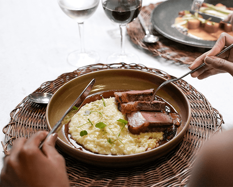 Prato com fatias de carnes e arroz, em prato de cerâmica, com mesa posta. Há taça de vinho ao fundo.