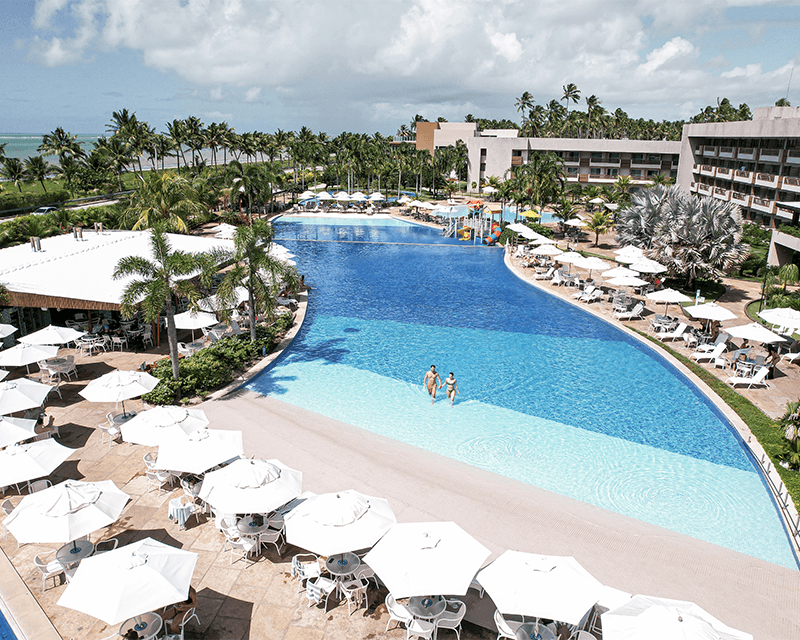 Vista aérea da piscina principal. O entorno é repleto de guarda-sóis e mesas brancas, coqueiros e o edifício principal do Japaratinga Lounge Resort. Ao fundo, linha de coqueiros na praia sob um céu parcialmente nublado.