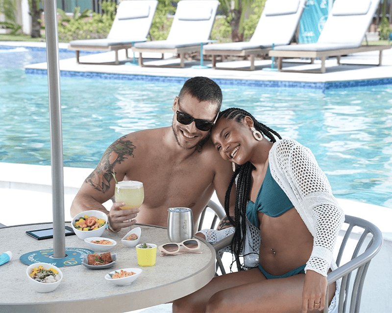 Casal sorrindo, sentados numa cadeira a beira da piscina. Na mesa tem petiscos e bebidas, álem de óculos escuros, protetor e um kindle. Ao fundo tem a piscina e espreguiçadeiras.