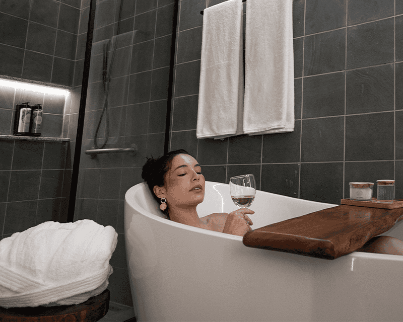 Uma mulher relaxando na banheira do Garden Exclusive, com uma taça de vinho branco em uma bandeja de madeira sobre a banheira. Ao lado há um chuveiro com box de vidro e paredes de azulejo cinza, e uma planta verde em um vaso.