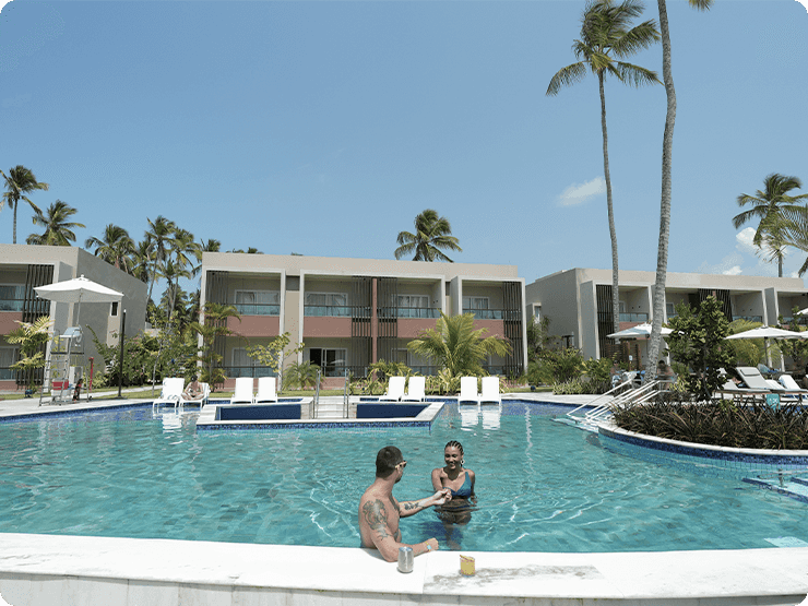Casal de mãos dadas na piscina do Gardens, com drinks na beira. O céu está sem nuvens e ensolarado.