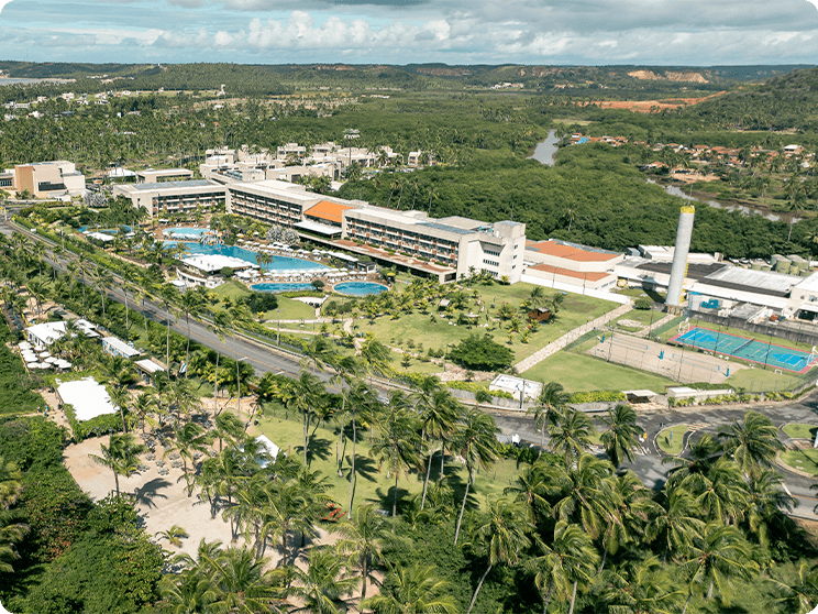 Vista aérea do Japaratinga Lounge Resort. É possível ve a área da piscina, quadras e gardens. Um pedaço da praia no canto com guarda-sóis brancos, espreguiçadeiras. Cercado por grande vegetação.