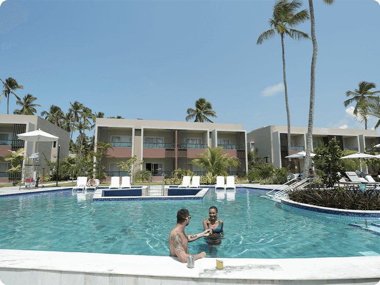 Casal de mãos dadas na piscina do Gardens, com drinks na beira. O céu está sem nuvens e ensolarado.