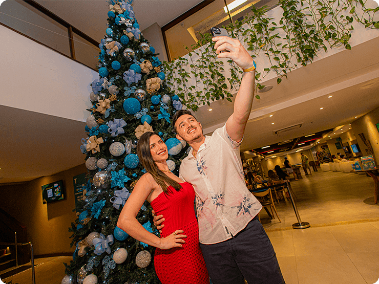 Casal tirando uma selfie em frente a grande árvore de natal na recepção do Japaratinga Lounge Resort, com a área de games ao fundo.