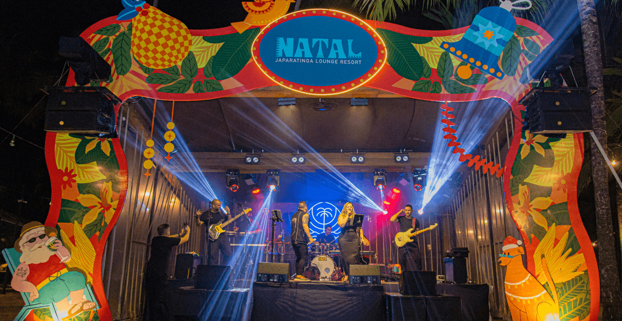 Palco do Natal no Japaratinga Lounge Resort. Decoração colorida de natal ao redor do palco, com uma banda de 6 integrantes cantando, vocalista sendo um homem e uma mulher. Há luzes coloridas no palco todo e coqueiros ao redor.