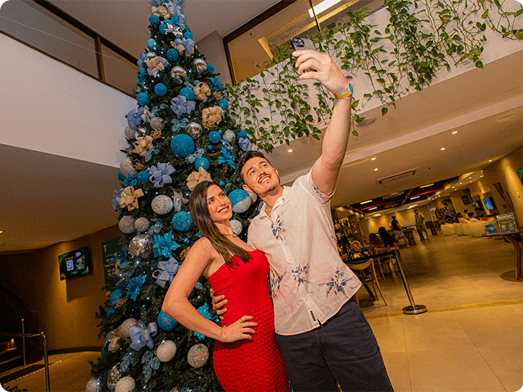 Casal tirando uma selfie em frente a grande árvore de natal na recepção do Japaratinga Lounge Resort, com a área de games ao fundo.