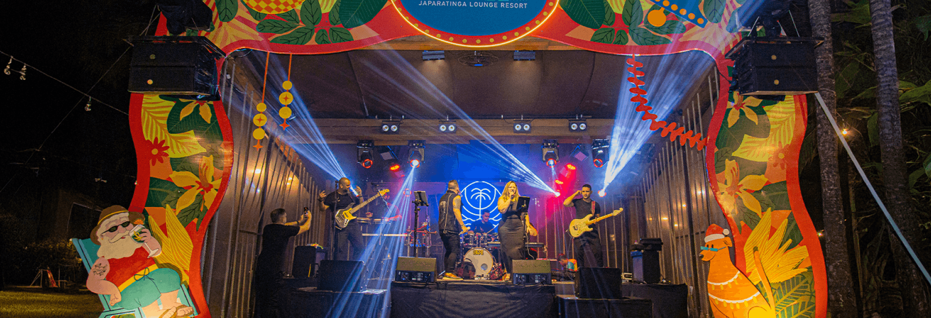 Palco do Natal no Japaratinga Lounge Resort. Decoração colorida de natal ao redor do palco, com uma banda de 6 integrantes cantando, vocalista sendo um homem e uma mulher. Há luzes coloridas no palco todo e coqueiros ao redor.