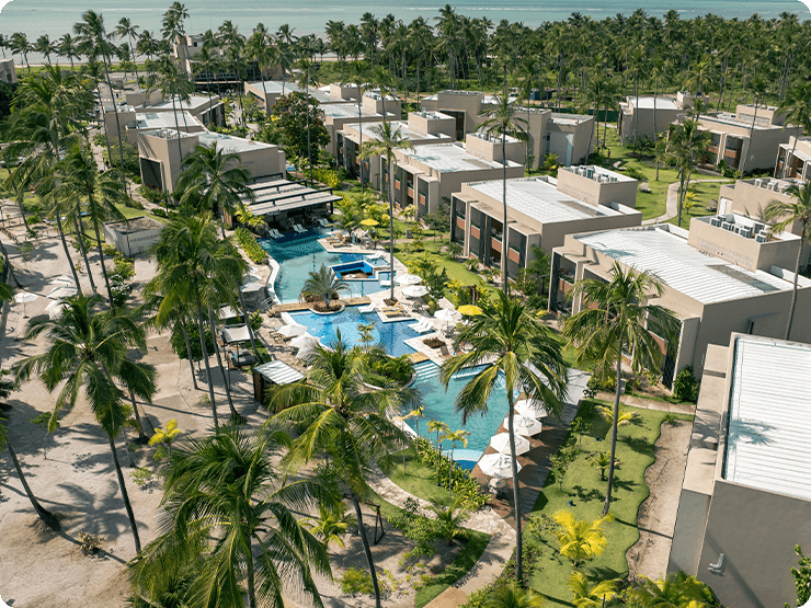 Vista aérea das Gardens Suítes do Japaratinga Lounge Resort, mostrando a área das piscinas com guarda-sóis brancos, espreguiçadeiras, cercadas por vegetação tropical e palmeiras