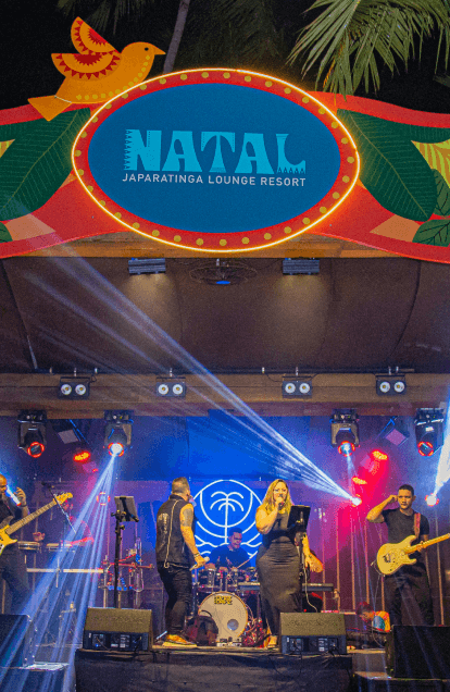 Palco do Natal no Japaratinga Lounge Resort. Decoração colorida de natal ao redor do palco, com uma banda de 6 integrantes cantando, vocalista sendo um homem e uma mulher. Há luzes coloridas no palco todo e coqueiros ao redor.